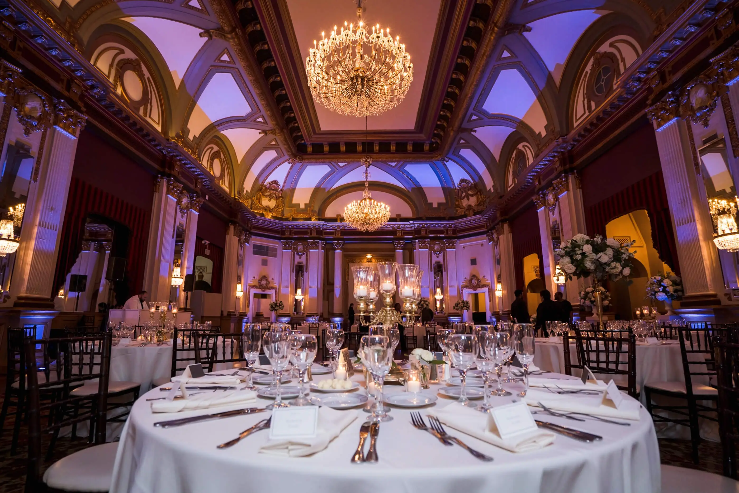 A ballroom set for dinner under chandeliers with purple uplighting