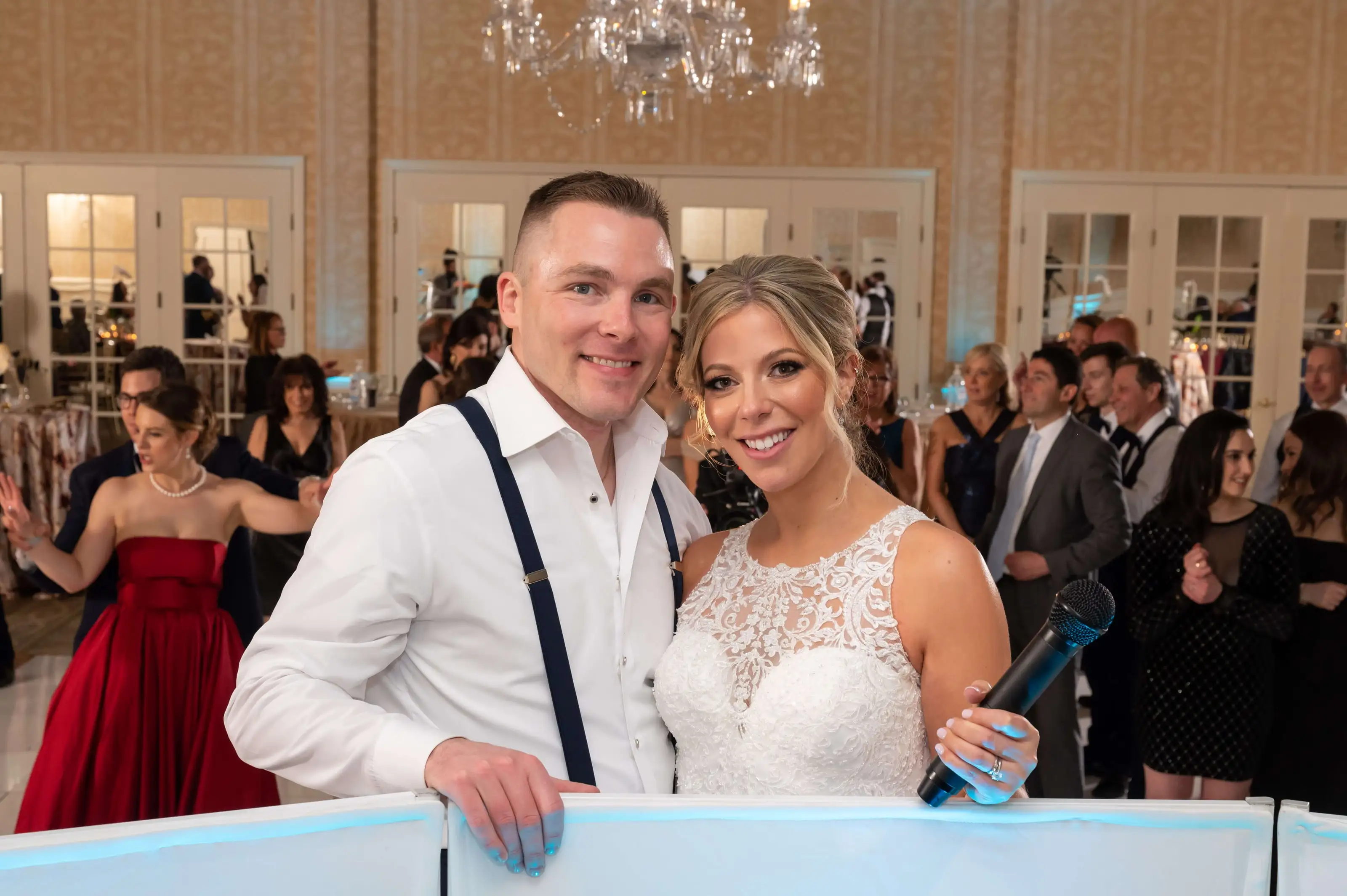 A bride and groom smiling at the DJ booth with their guests dancing behind them