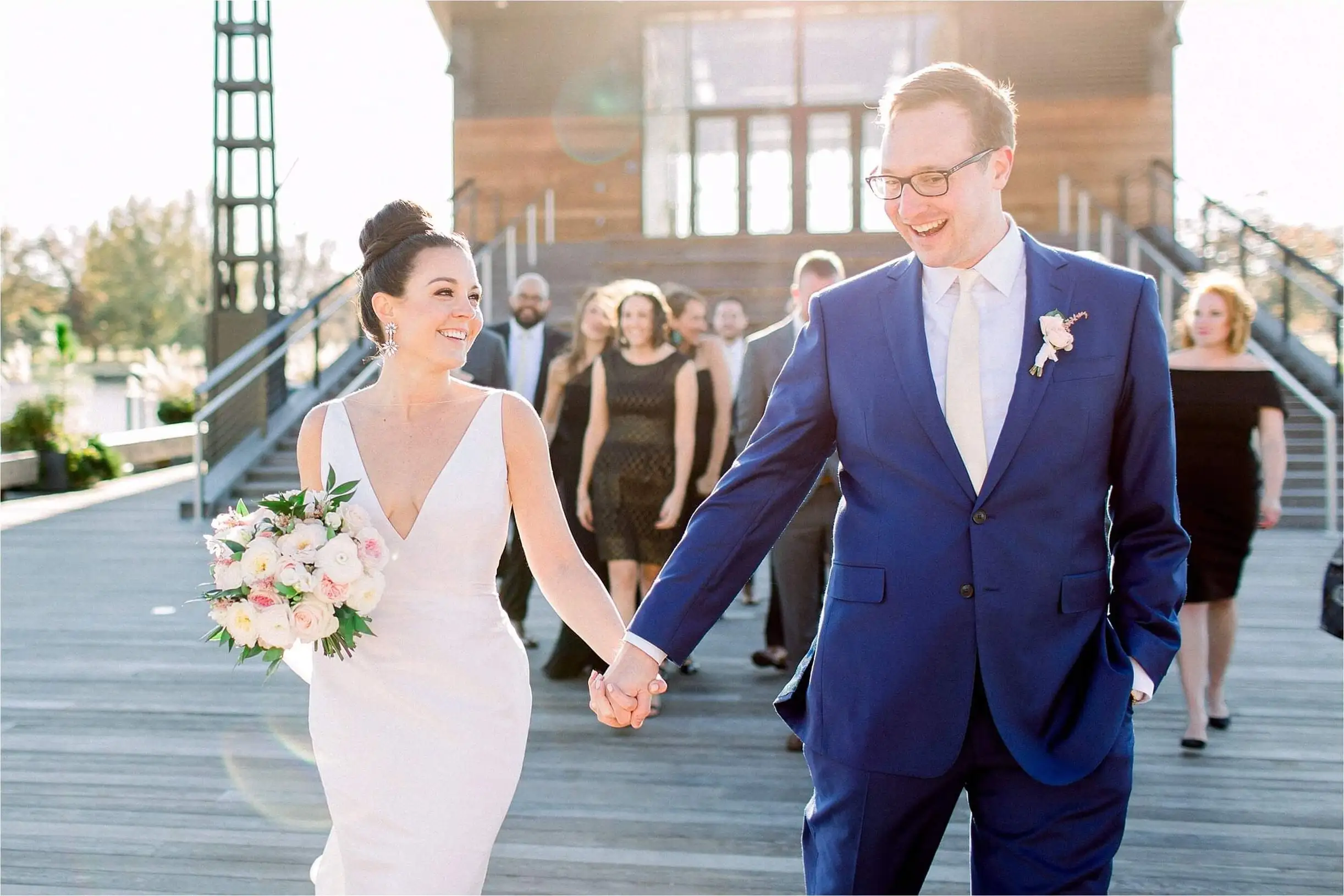 A bride and groom walking hand in hand along a boardwalk with their guests behind them