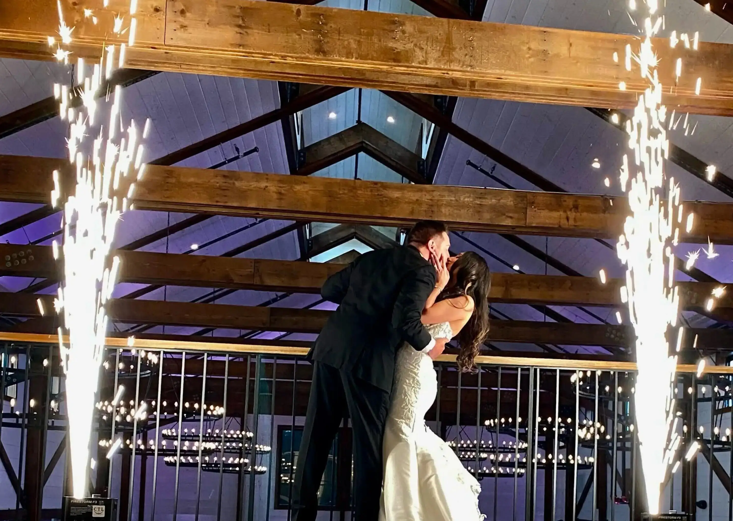 A bride and groom kissing on the dance floor between two fountains of sparks under wooden beams