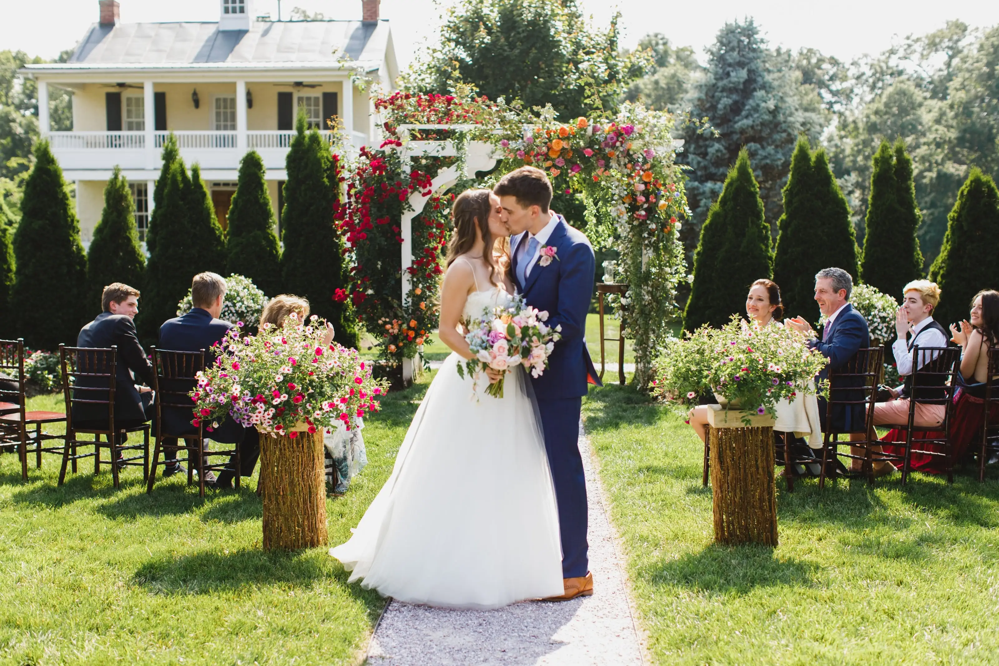 A bride and groom kissing at the end of a garden aisle lined with flowers