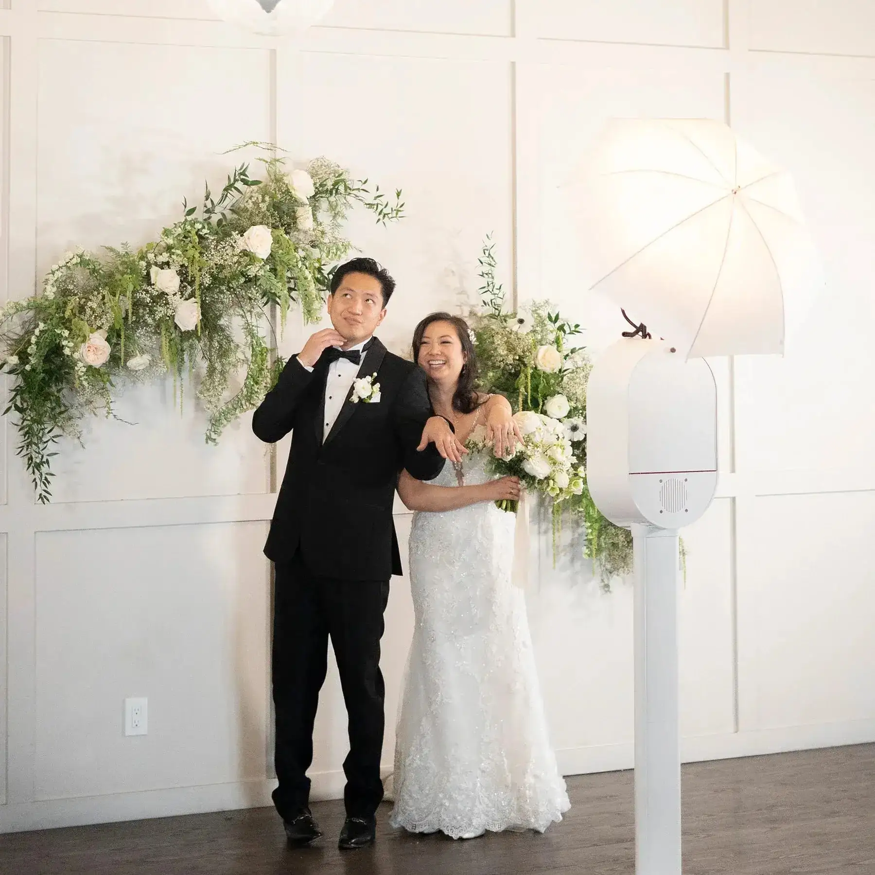 A bride and groom posing at a photo booth in front of a flower wall