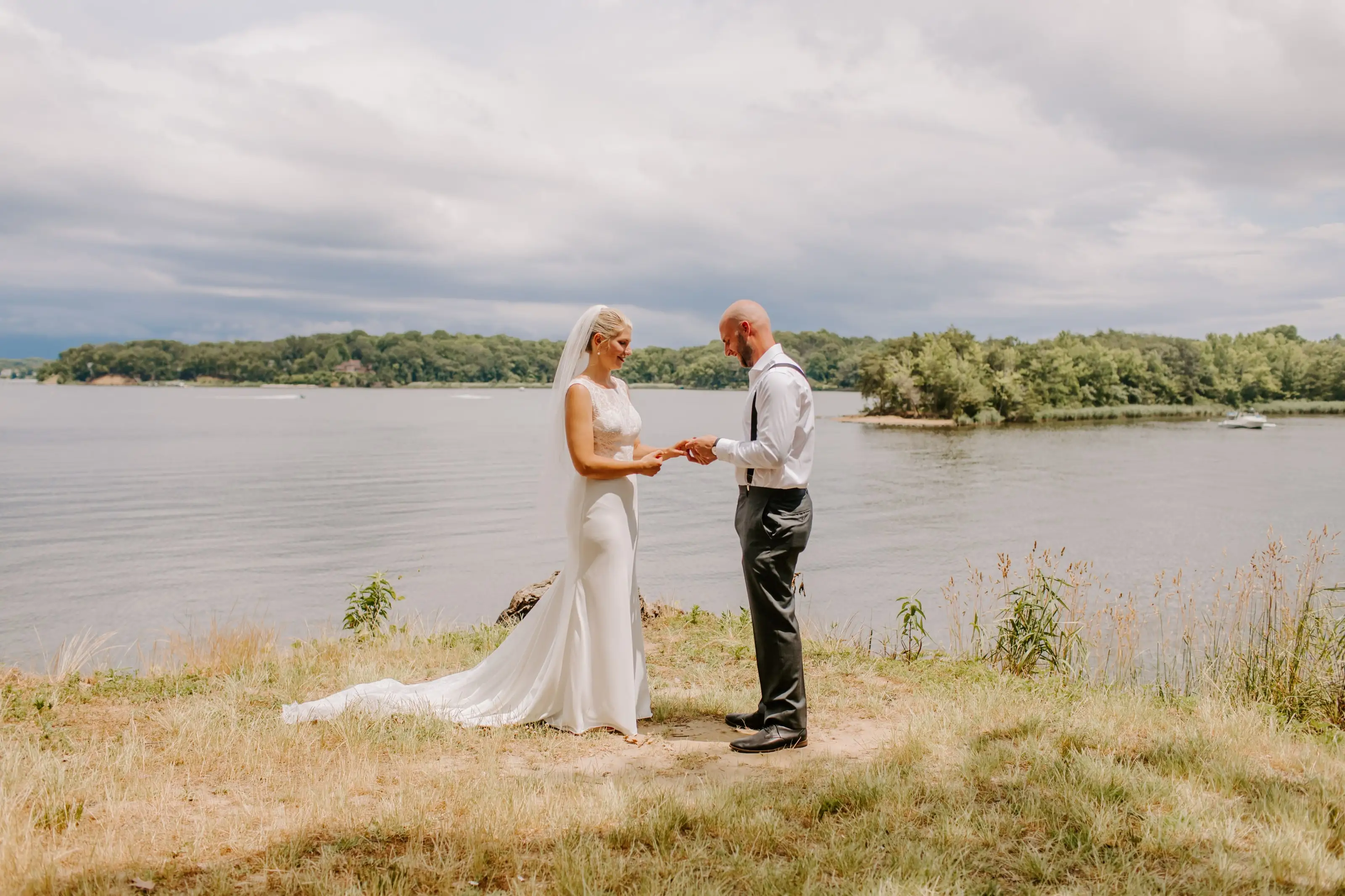 A bride and groom exchanging rings on a grassy bank above the water
